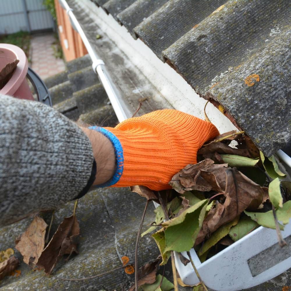 A person wearing orange gloves is cleaning a gutter with leaves