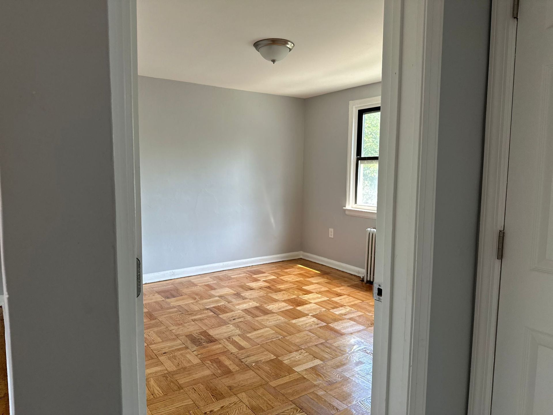 View through an open doorway into an empty room with gray walls, parquet floors, and a window.