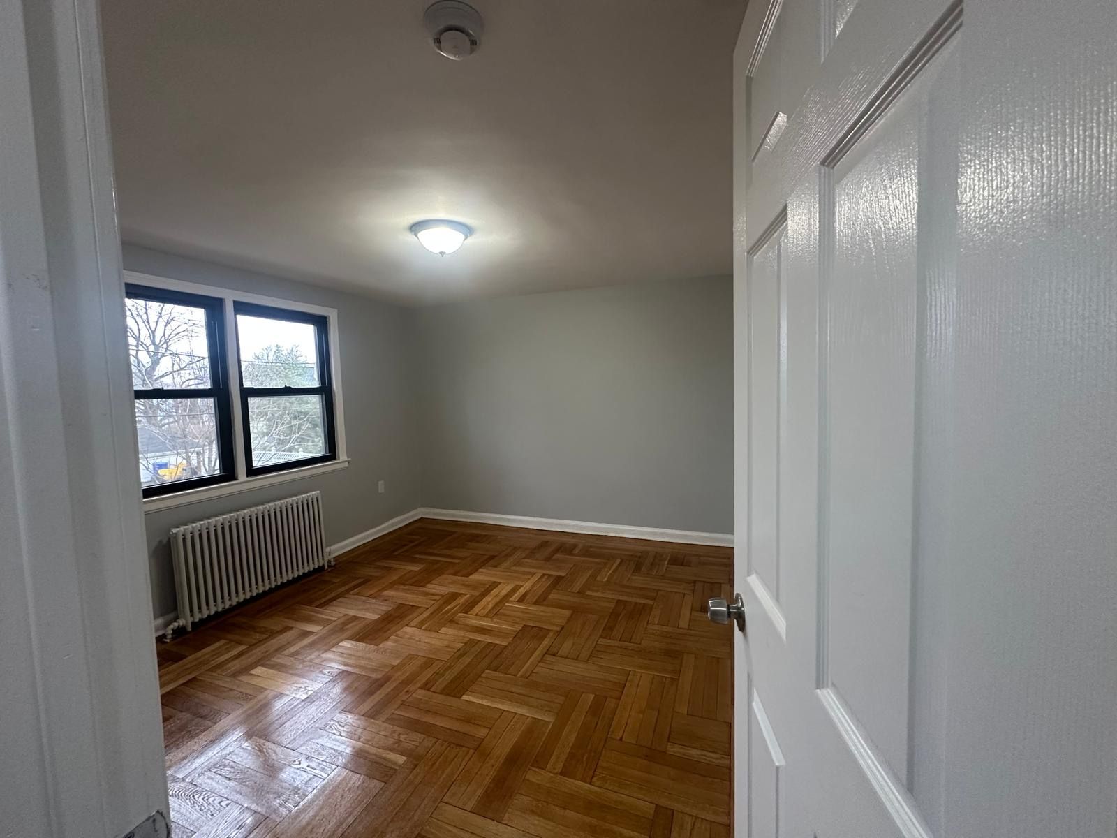 Empty bedroom with parquet floors, window, radiator, and gray walls, viewed from an open door.