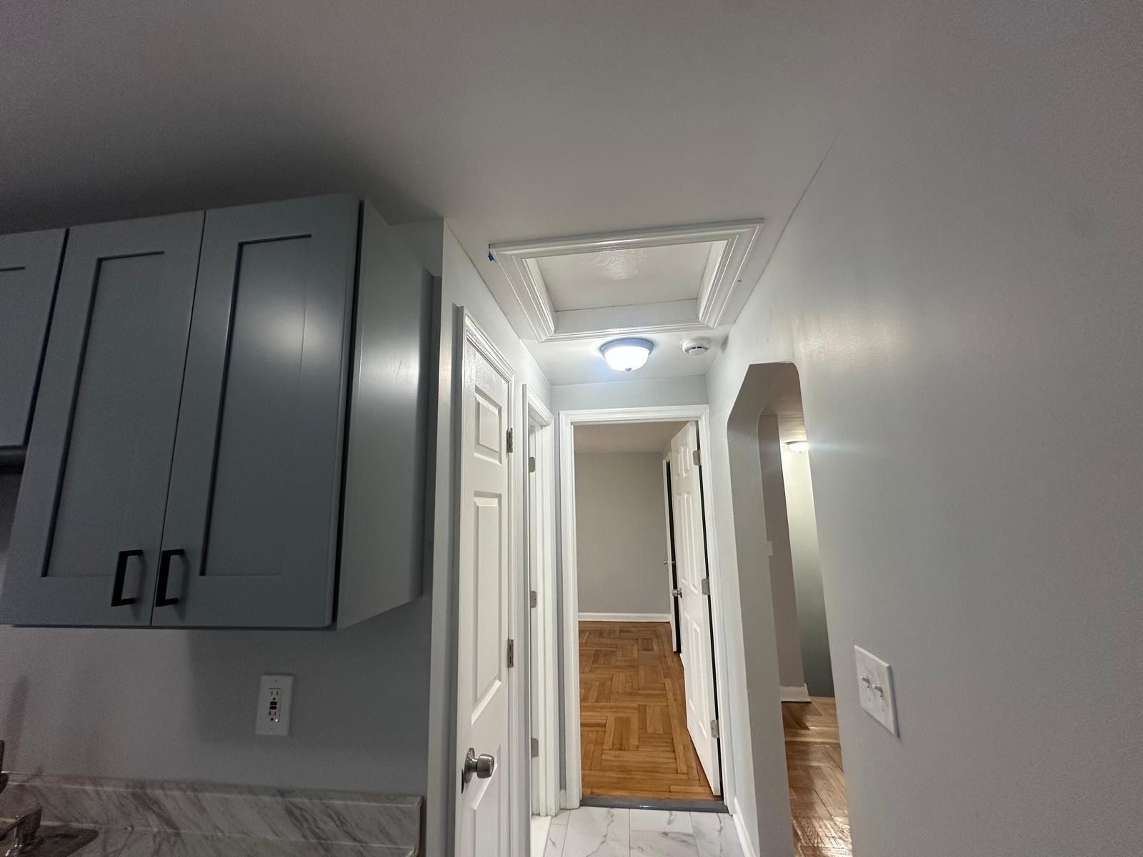 View down a hallway with white doors, gray walls, and an open ceiling hatch; cabinets on the left.