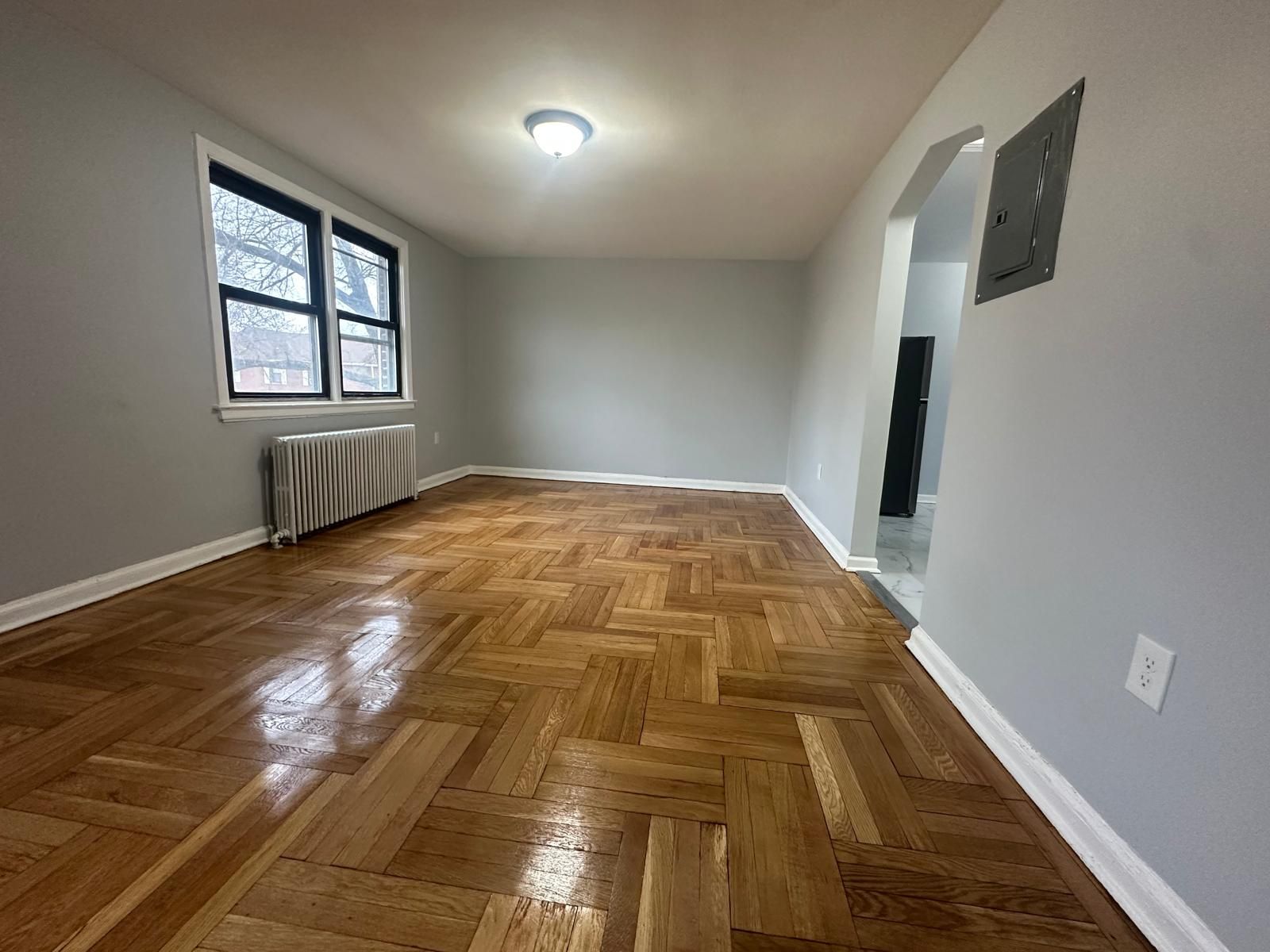 Empty room with hardwood floors, window, radiator, and doorway. Gray walls.