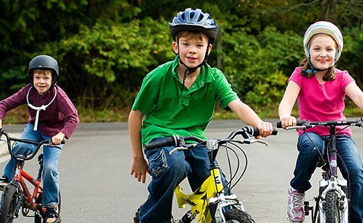 Three children in helmets riding bicycles on a paved road lined with trees.