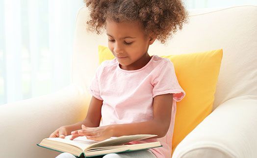 A person sitting in a white armchair against a yellow cushion, focused on reading an open book.