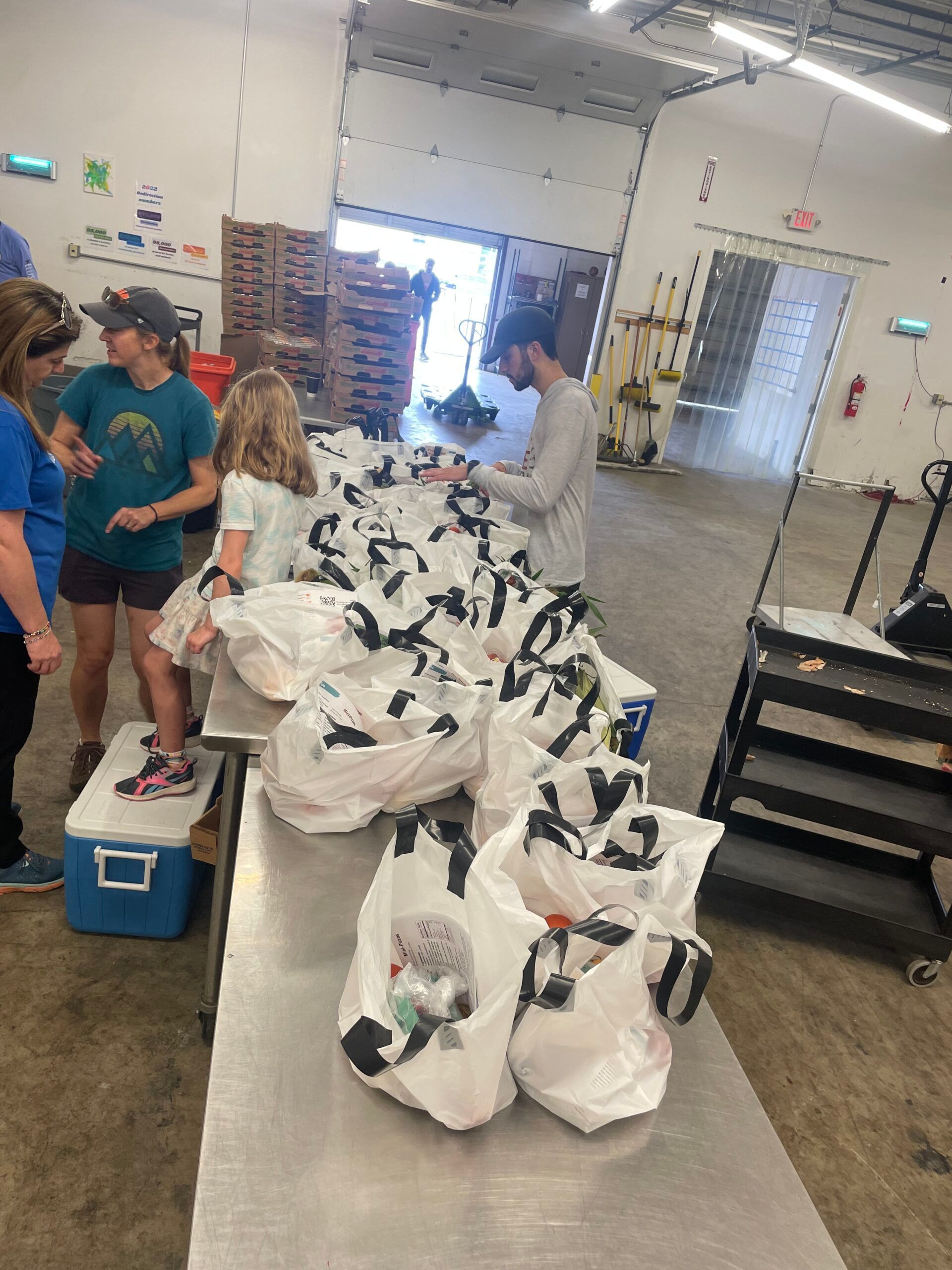 Three people stand around a metal table packing white bags for distribution in a large warehouse space.