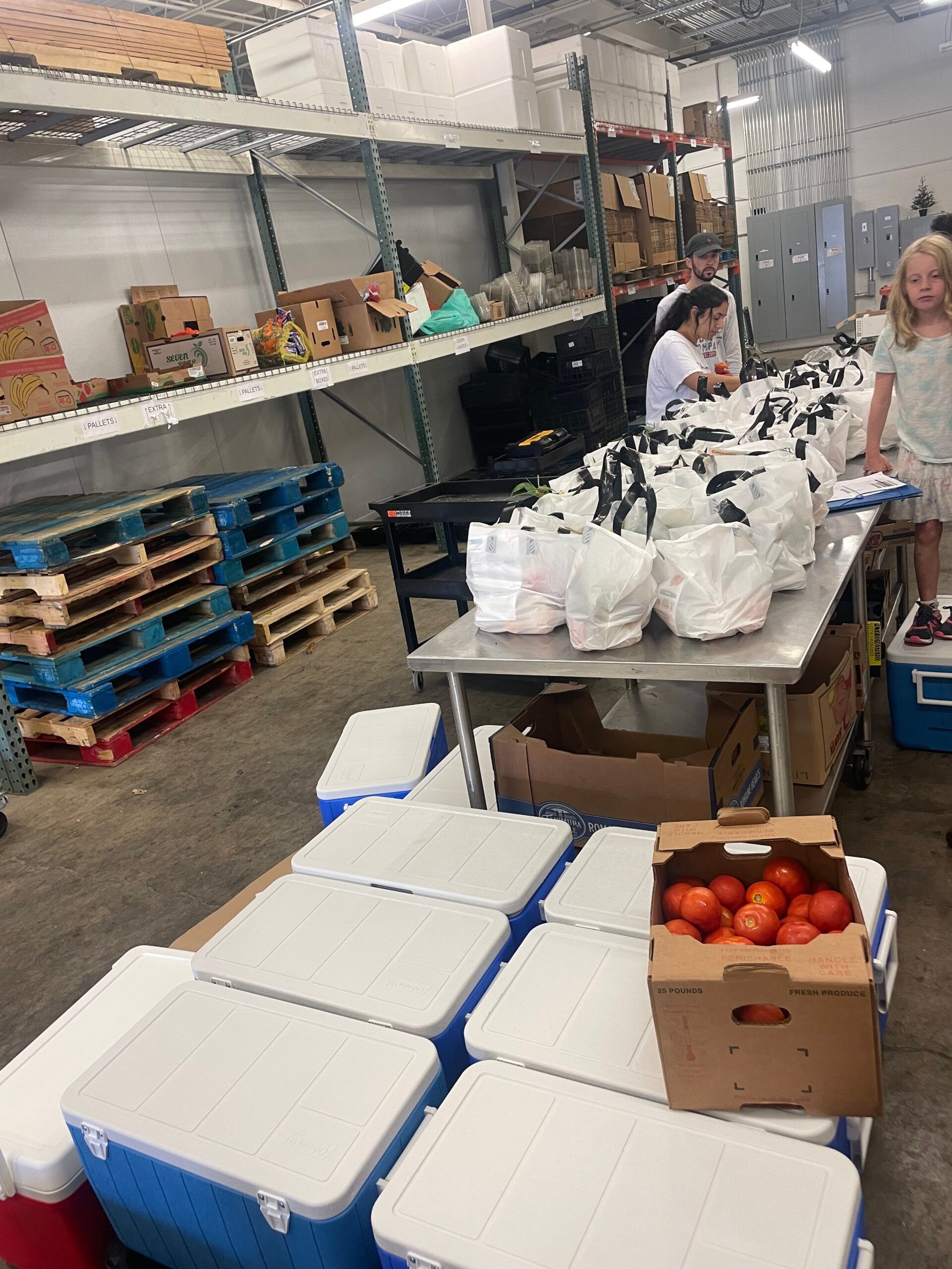 Volunteers prepare bags of food on a table in a warehouse setting filled with pallets and cooling chests.