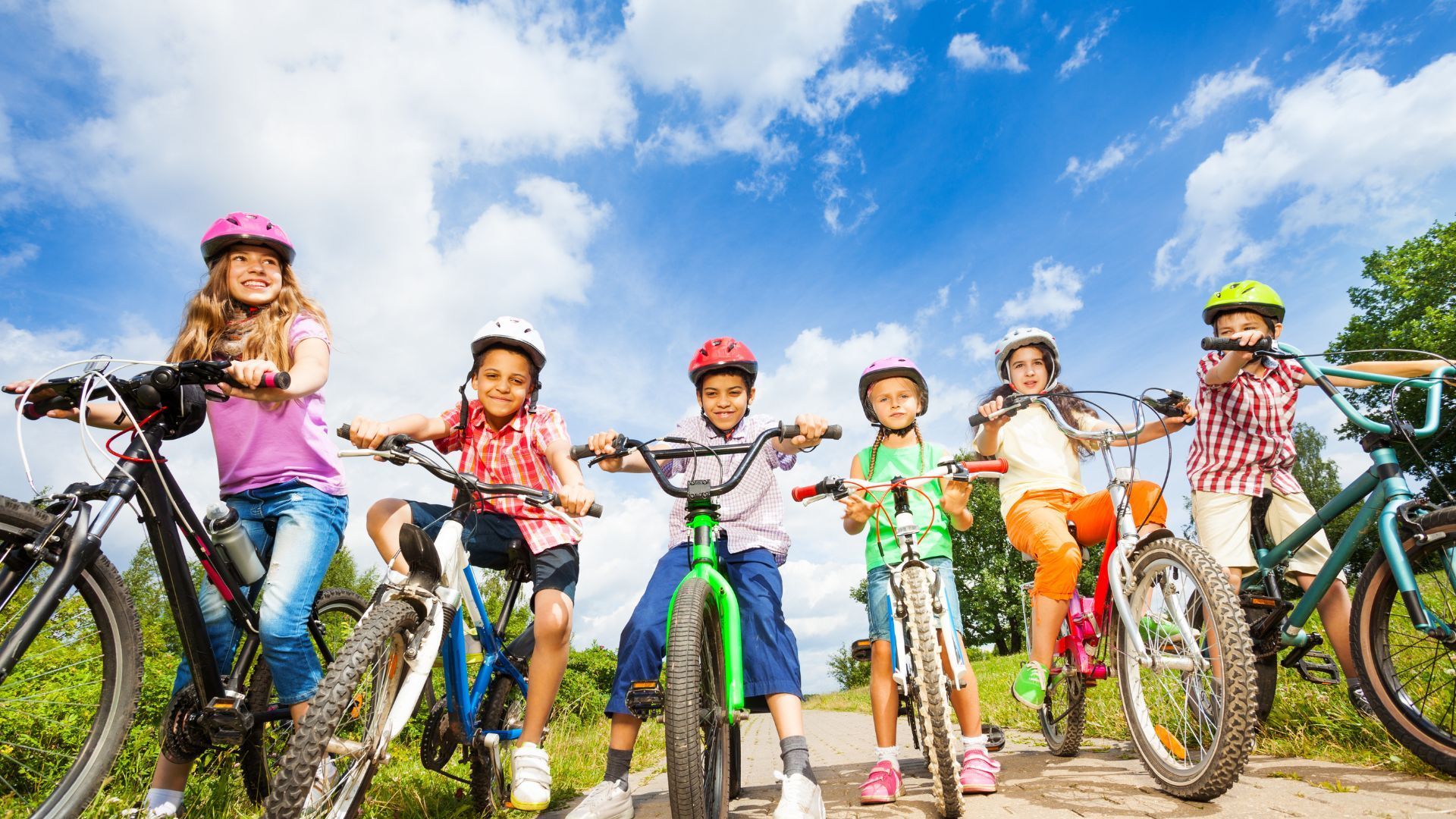 Six children in helmets sit on bicycles on a dirt path under a bright, sunny blue sky.