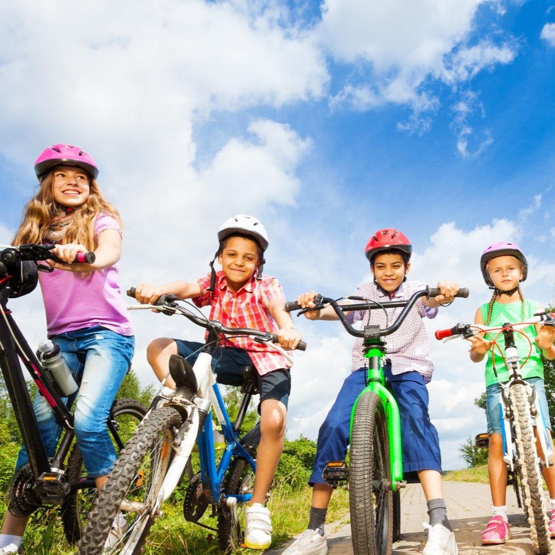 Four children wearing helmets smile while riding bicycles on a paved path against a bright blue sky.