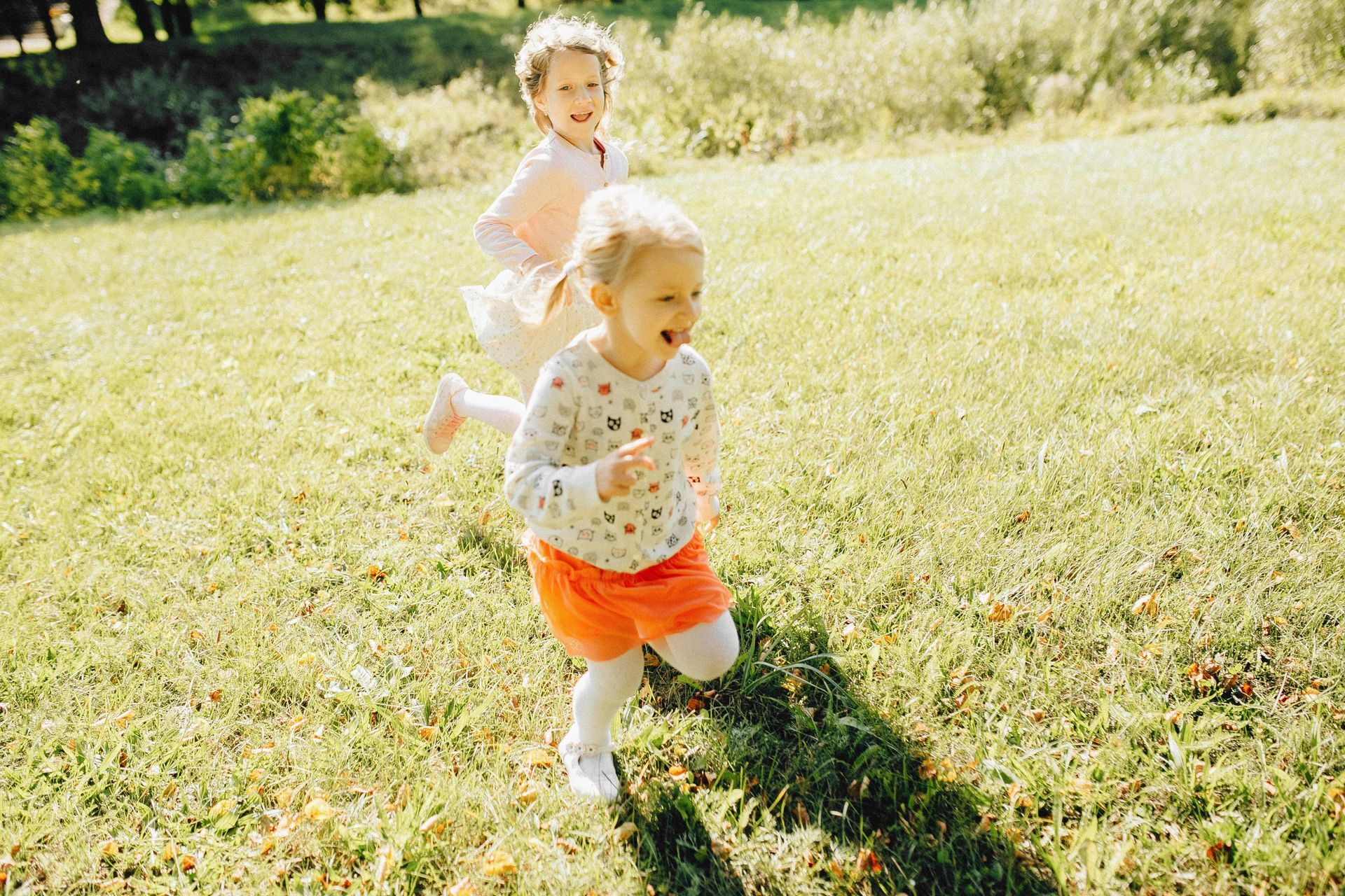 Two children run across a sunny, grassy field, both smiling and looking ahead.