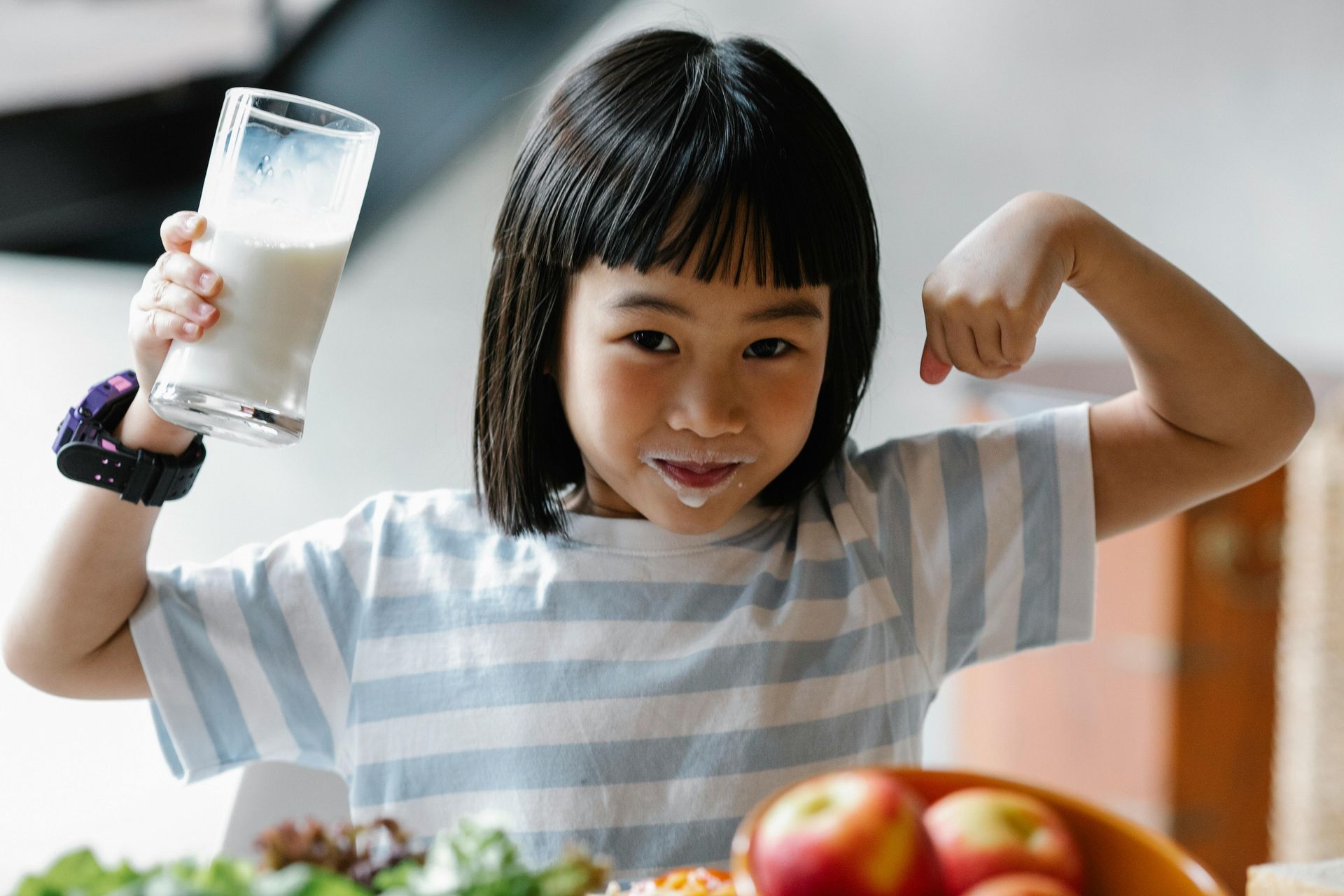A child in a striped shirt flexes an arm and holds a glass of milk with a milk mustache, near a bowl of fruit.