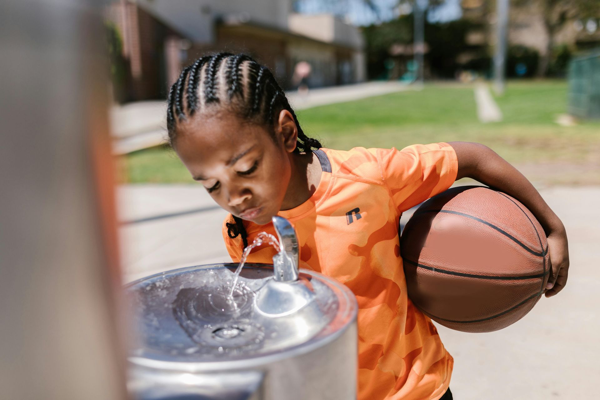 A child in an orange shirt drinks from an outdoor water fountain while holding a basketball.