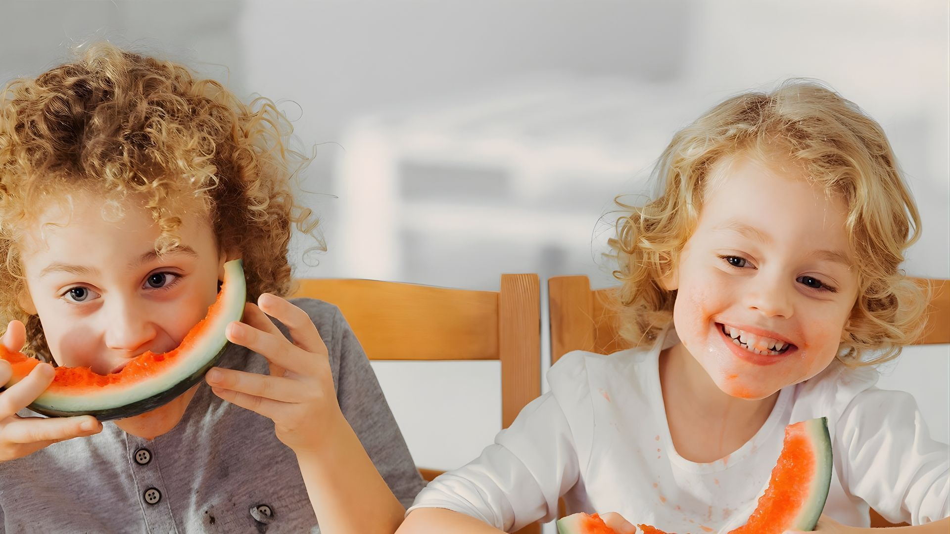 Two smiling children with curly hair eating slices of watermelon at a wooden table.