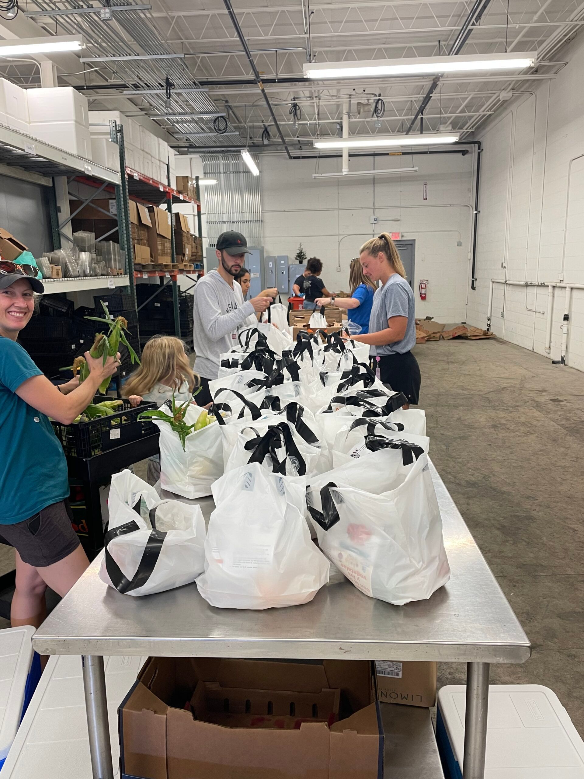 Volunteers pack food bags at a long metal table inside a warehouse.