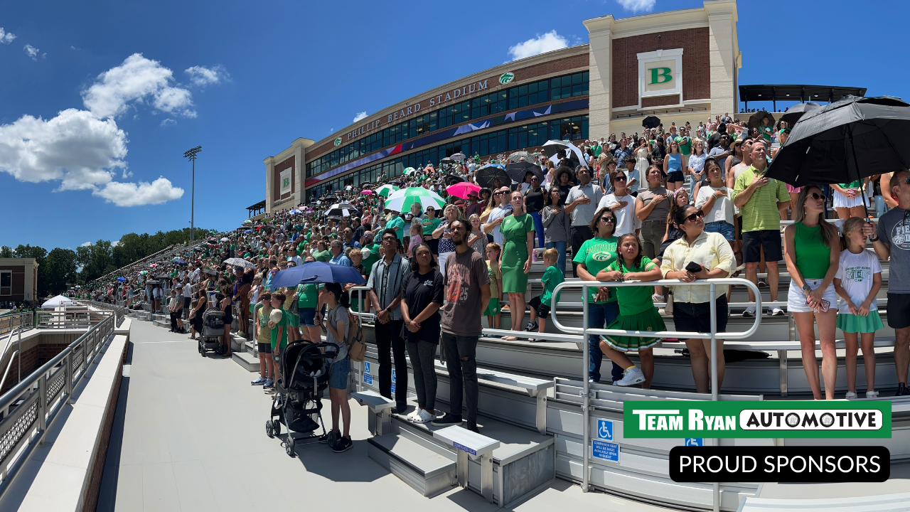 Fans in green crowd a stadium on a sunny day. Team colors and sponsor logo visible.