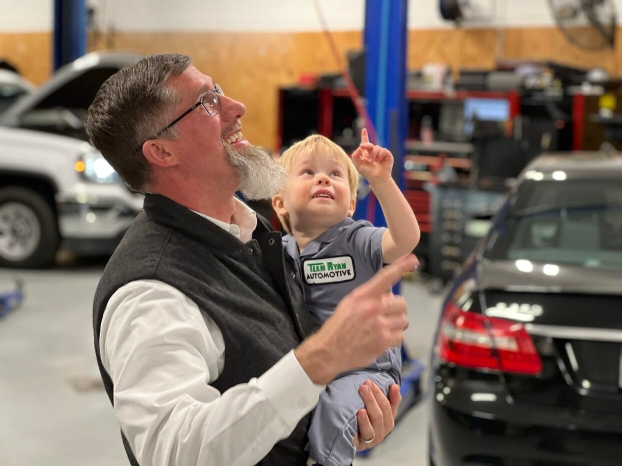 Man holding a child, both pointing up in a car repair shop. They are smiling.