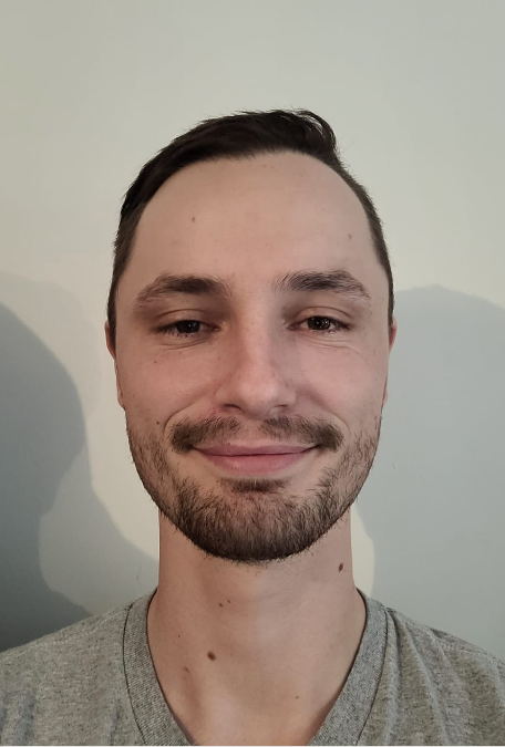 Un homme barbu sourit à la caméra tout en portant une chemise grise.