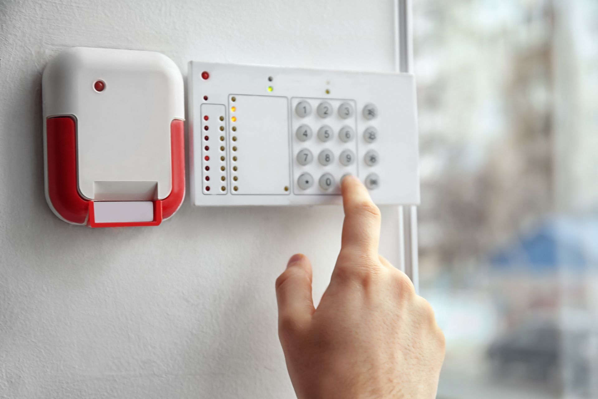 A hand pressing a keypad on a wall-mounted home security alarm panel beside a red siren unit