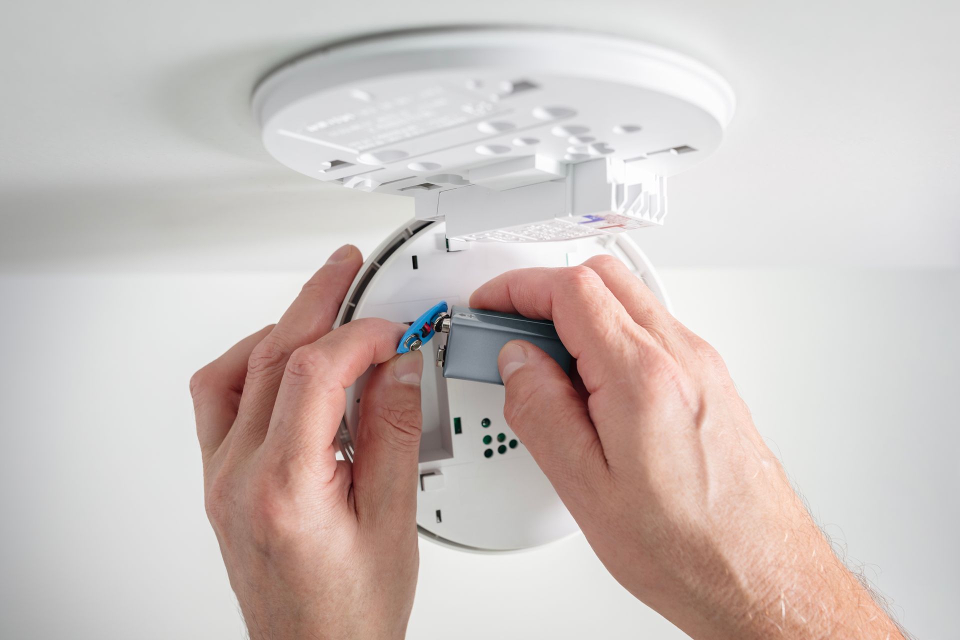 Hands installing a battery into a white smoke detector attached to a ceiling. Hands installing a battery into a white smoke detector attached to a ceiling.