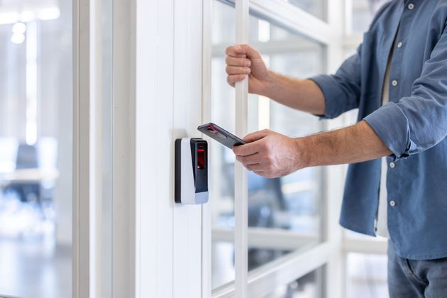 Man unlocking a glass door with a smartphone near a mounted access control device.