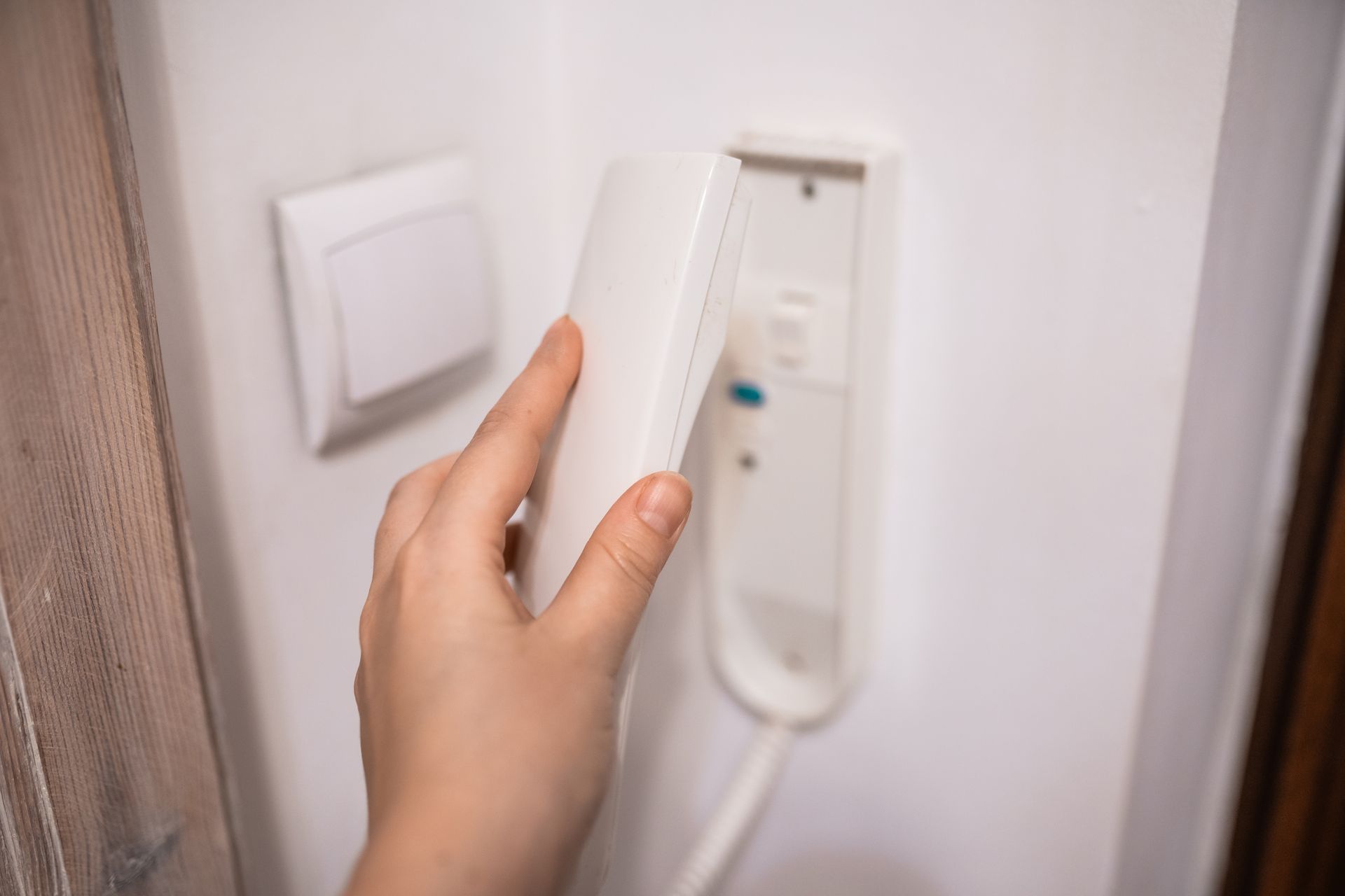 A hand reaching for a white intercom phone mounted on a wall. A white light switch is to the left.