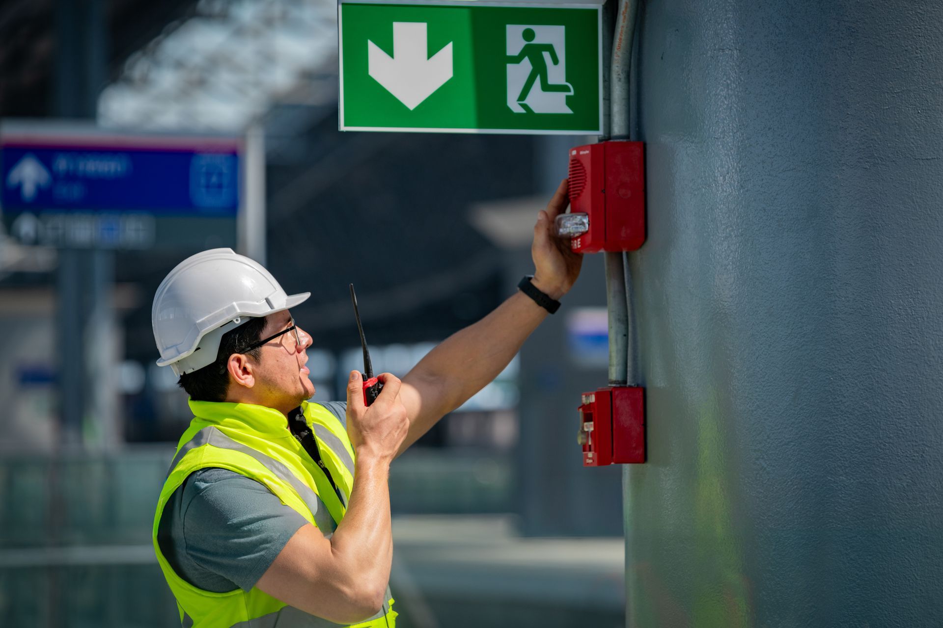 Construction worker in hard hat and vest testing fire alarm near exit sign. Construction worker in hard hat and vest testing fire alarm near exit sign.