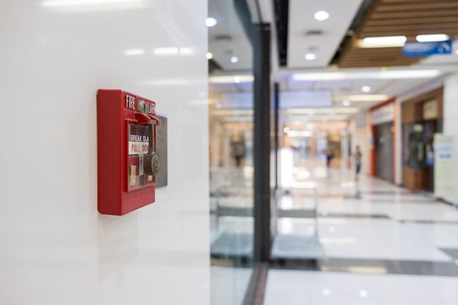 Red fire alarm box mounted on a white wall in a shopping mall hallway.