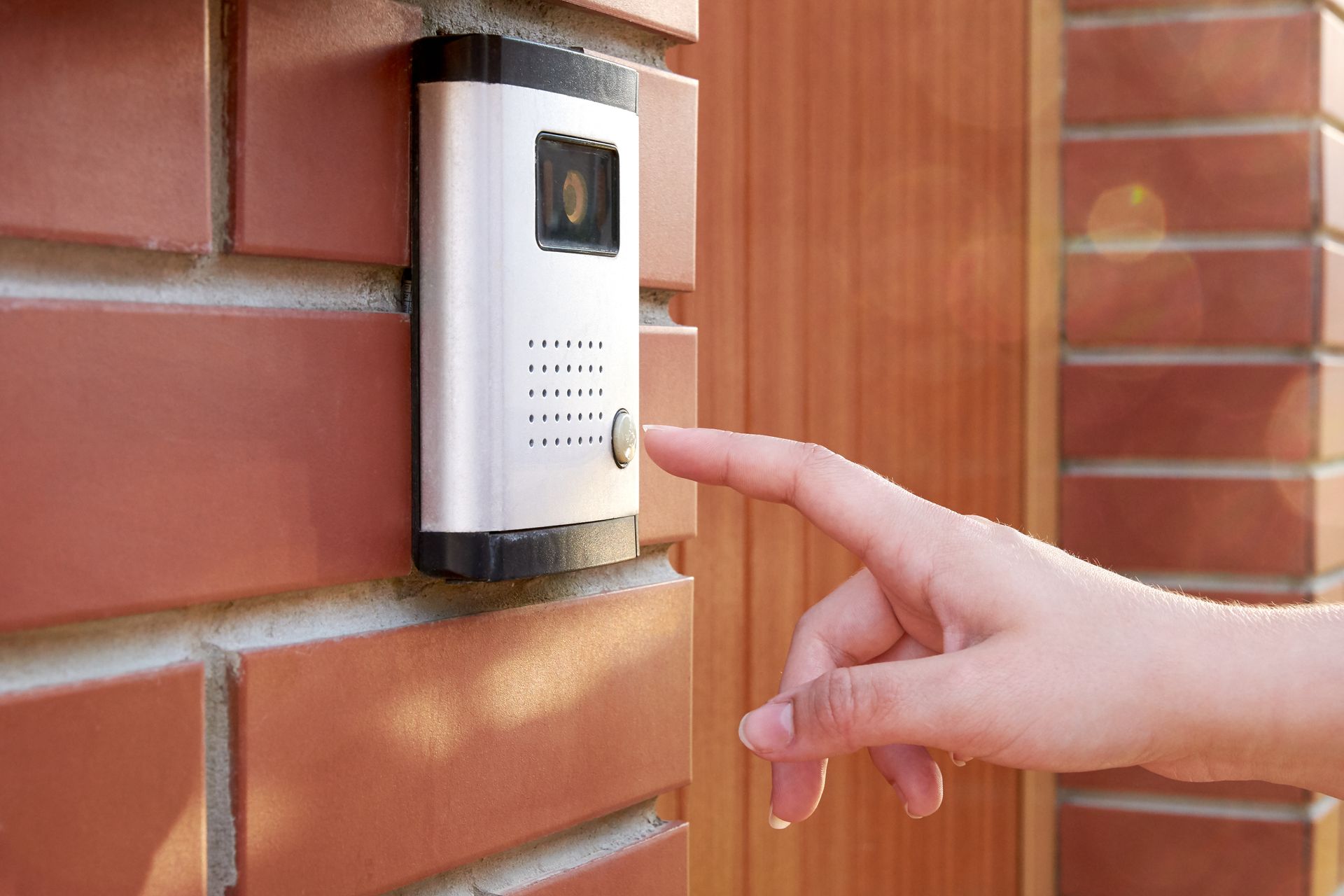Hand pressing the button on a silver doorbell with a camera, mounted on a brick wall.