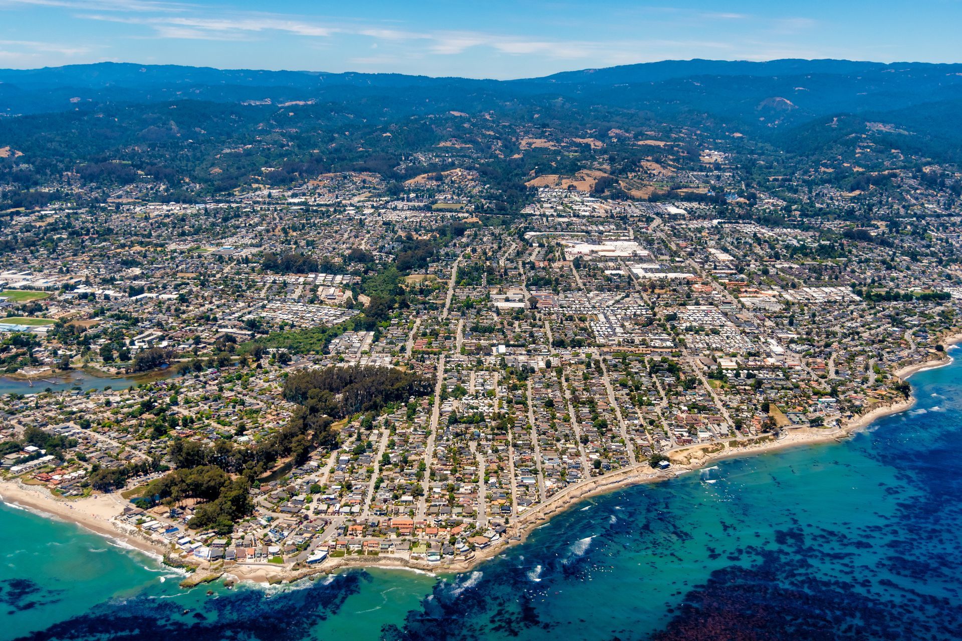 Aerial view of a coastal town with houses, roads, and a clear blue ocean under a sunny sky.