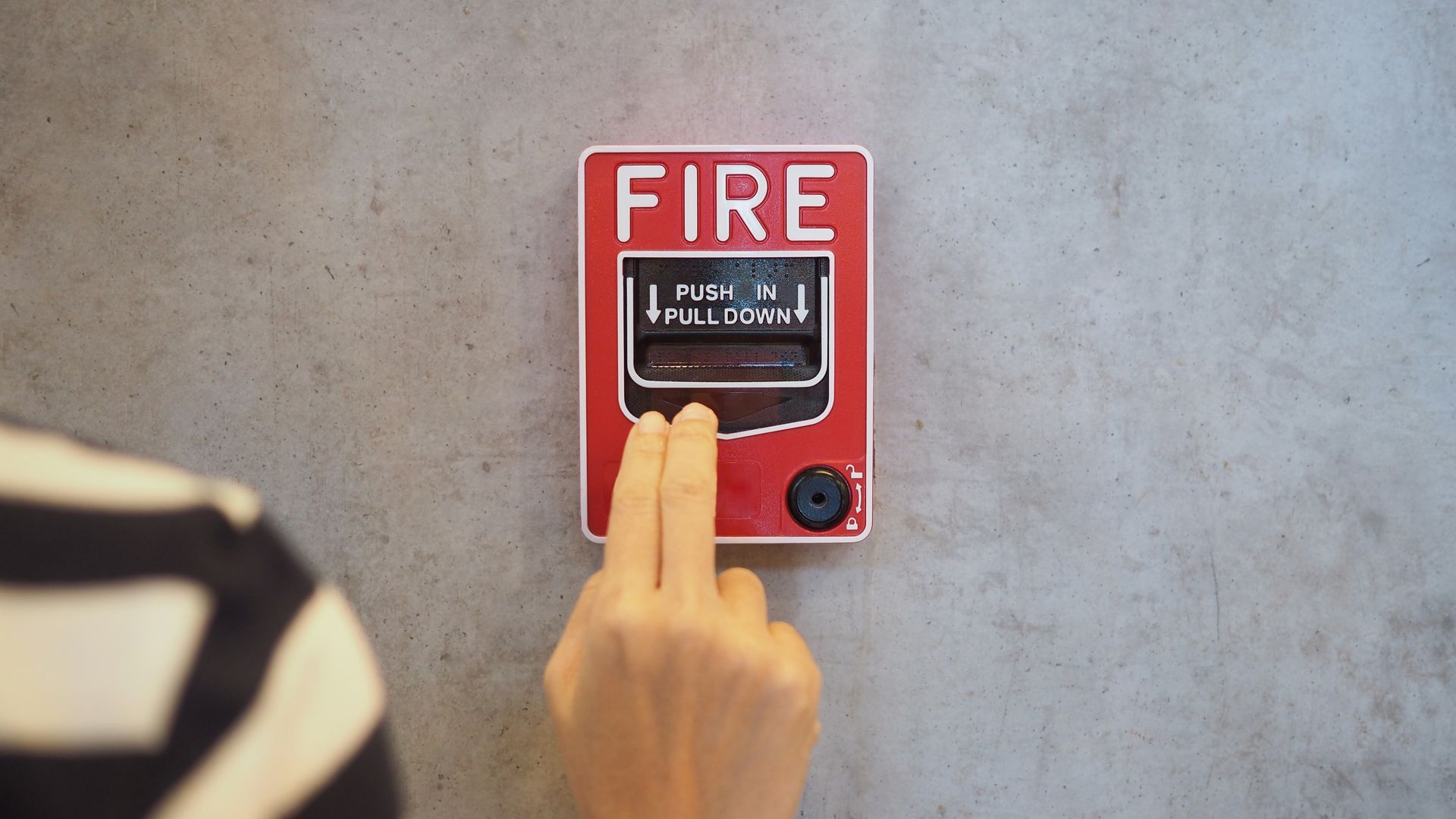 Hand pulling the lever on a red fire alarm box mounted on a gray wall. Hand pulling the lever on a red fire alarm box mounted on a gray wall.