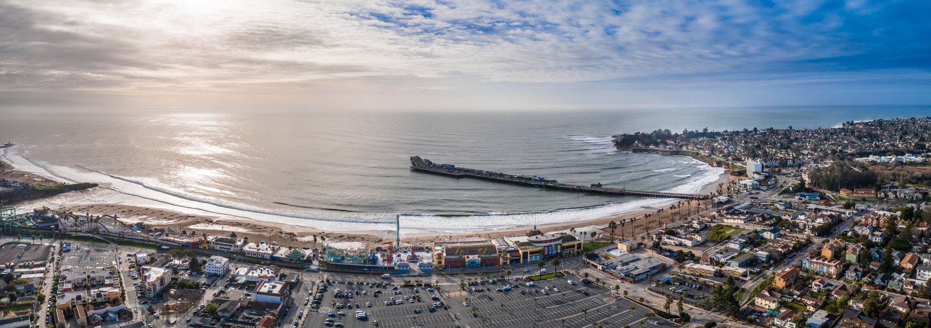 Panoramic aerial view of a beach town with a pier, ocean, beach, buildings, and a cloudy blue sky.