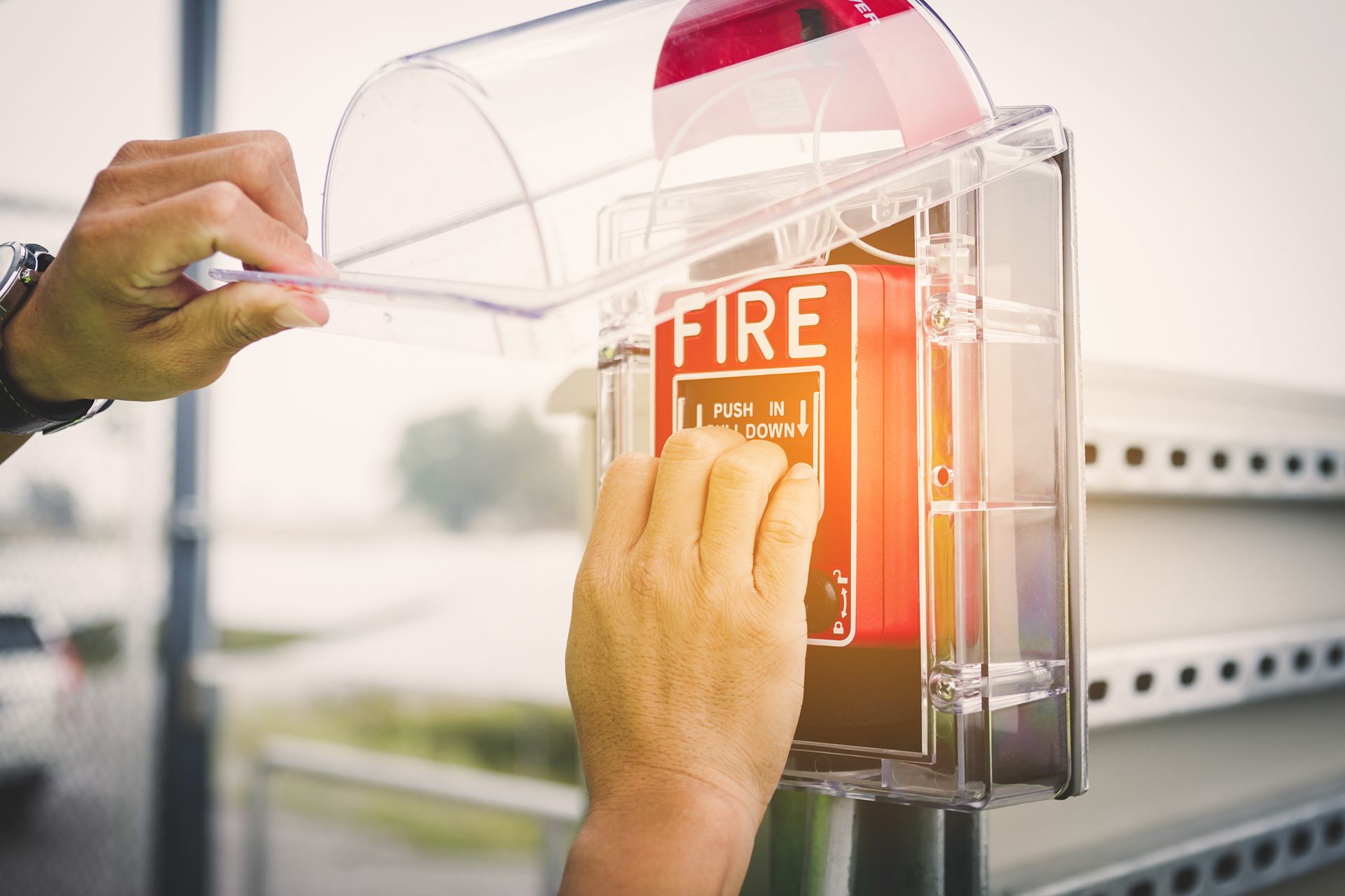Person activating a fire alarm, opening protective cover. Bright red fire alarm panel with clear plastic cover.