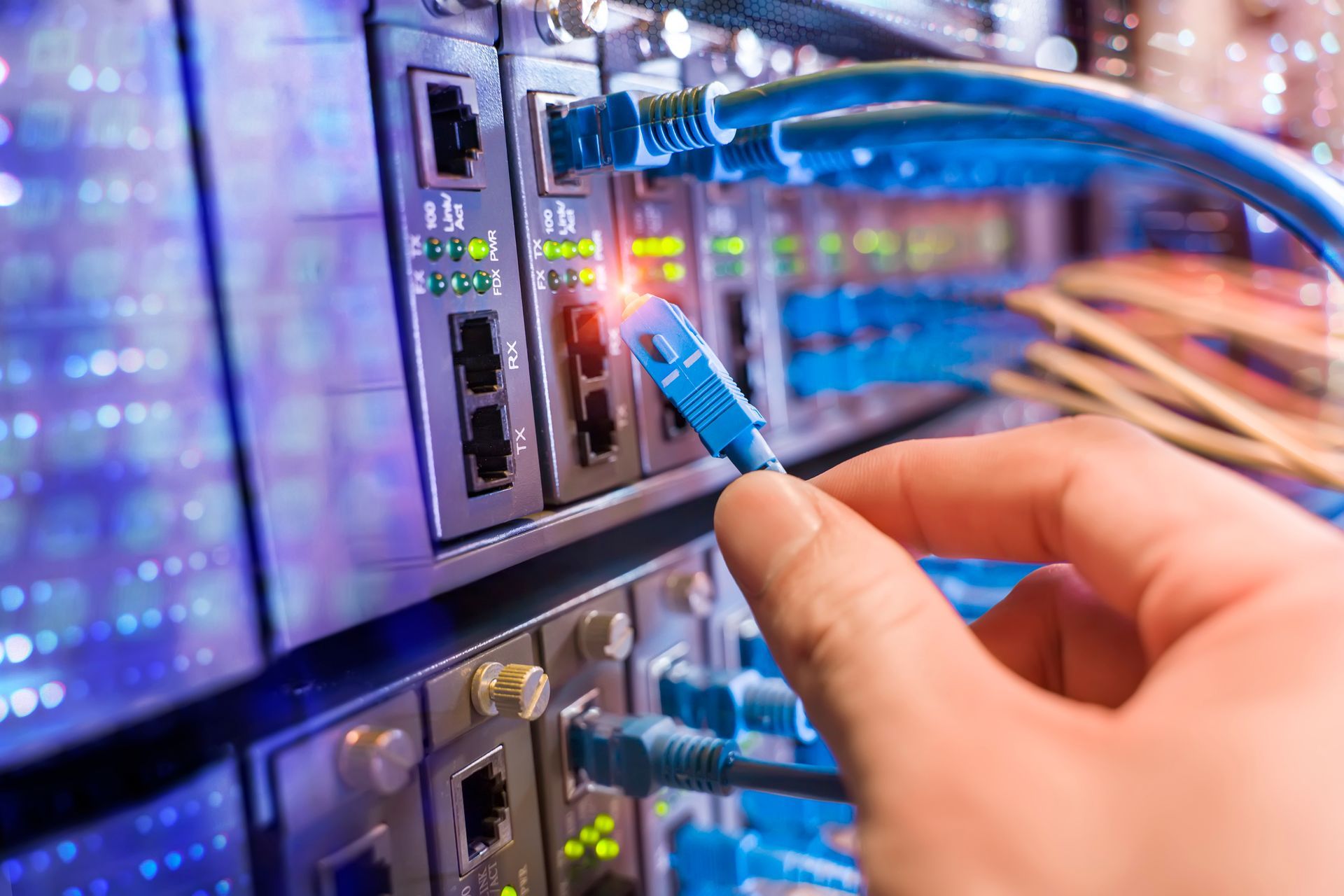 Hand plugging a blue Ethernet cable into a server rack.  Bright blue cables and lights against a metal background.