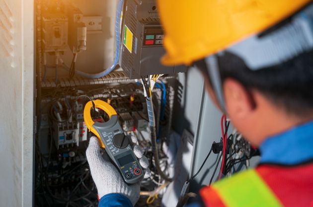 Electrician using a clamp meter on electrical panel. Yellow helmet, gloves, wires visible.