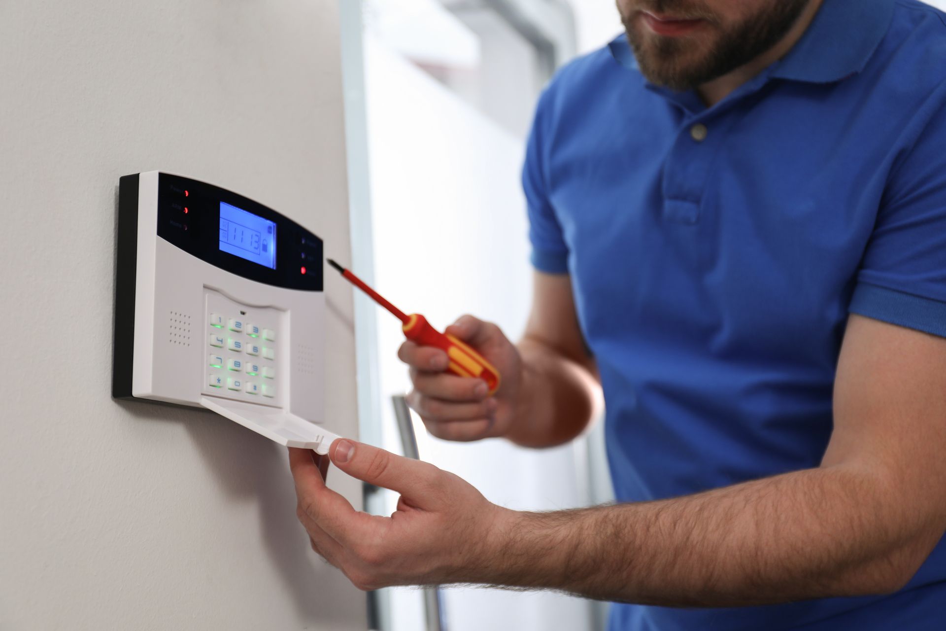 Person in blue shirt installing a security alarm system on a white wall with a screwdriver.
