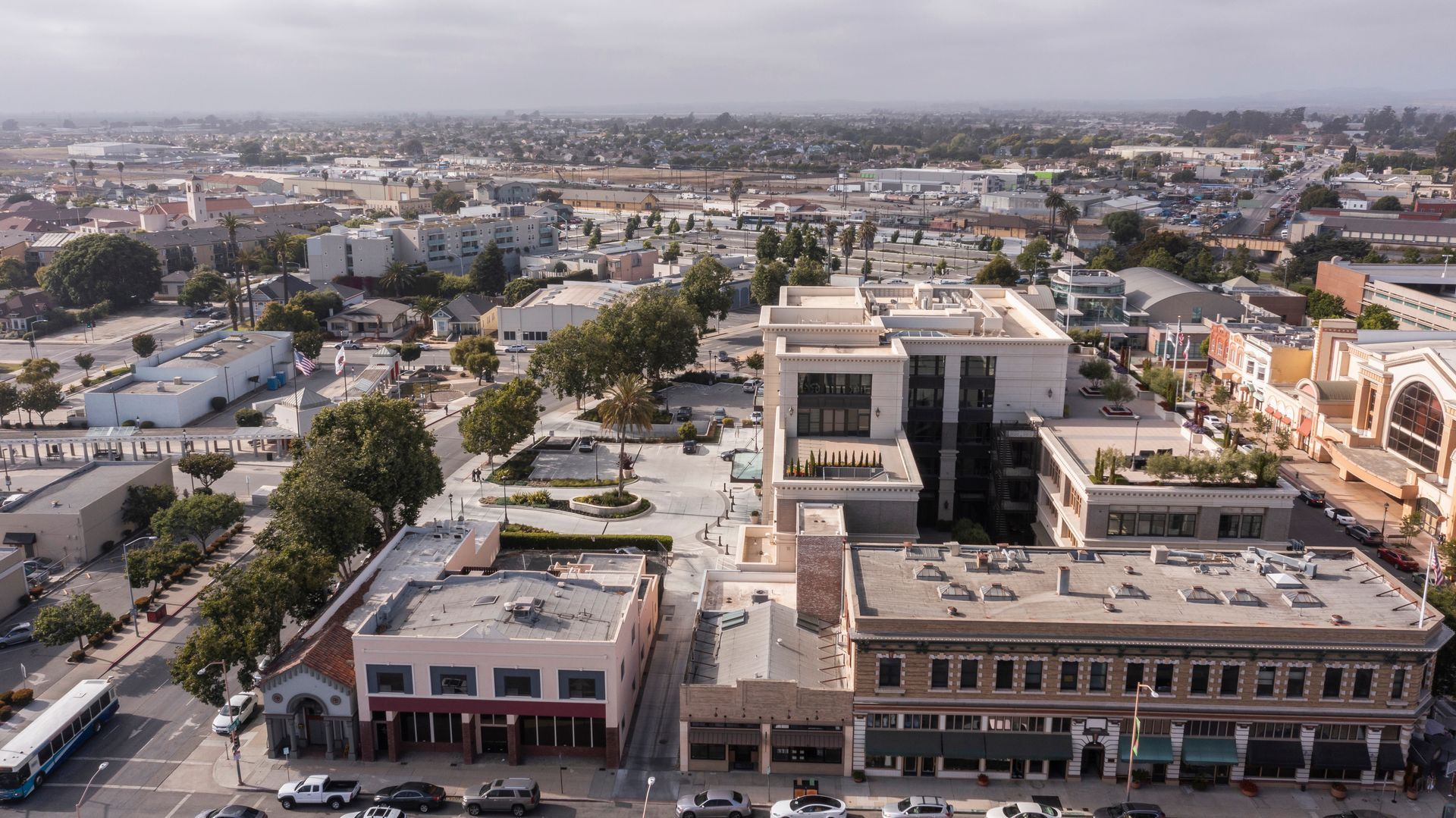 Aerial view of a town square with buildings, trees, and parked cars, under a cloudy sky.