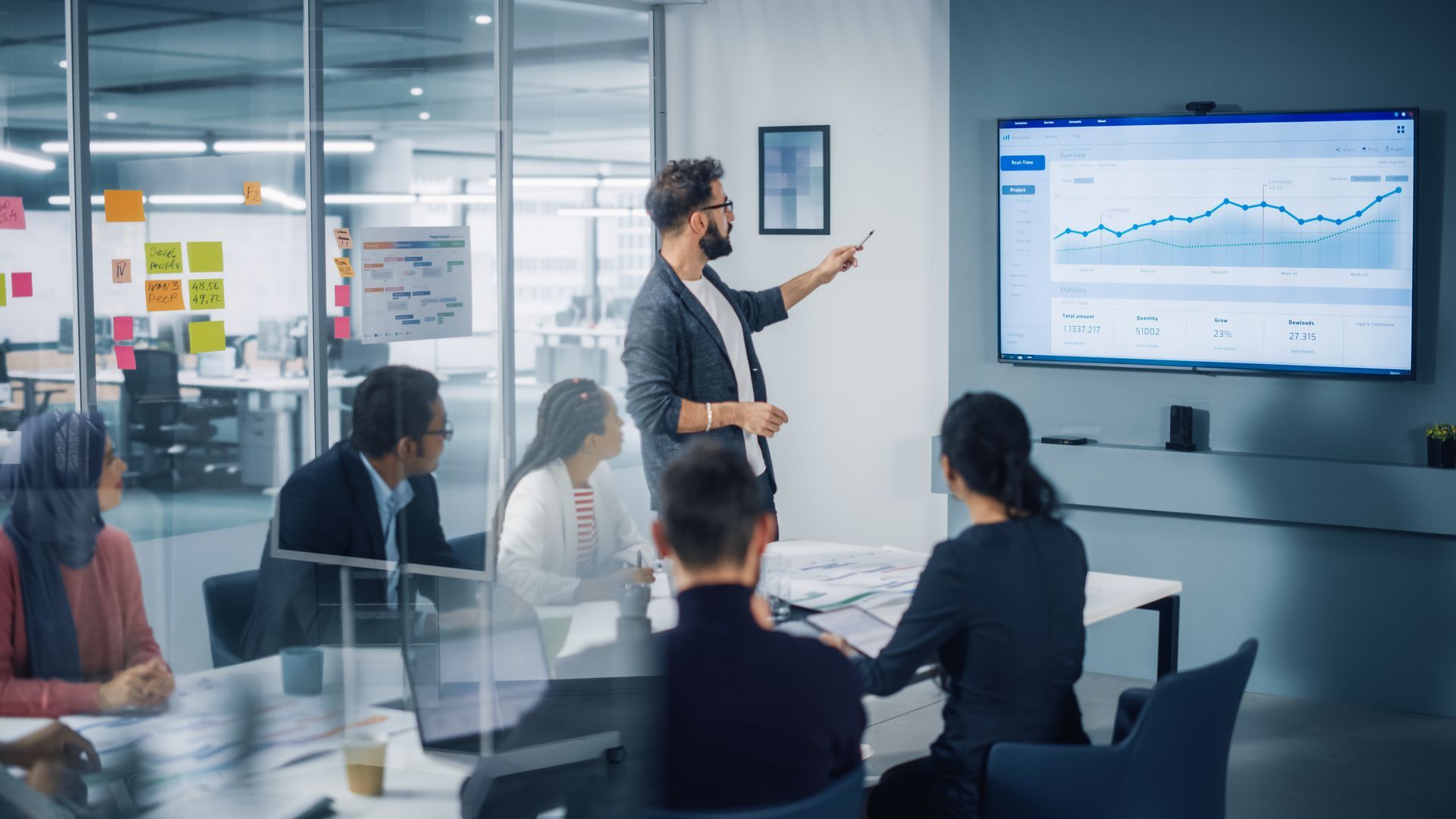 Man presenting data to colleagues in a modern office, pointing at a graph on a large screen.