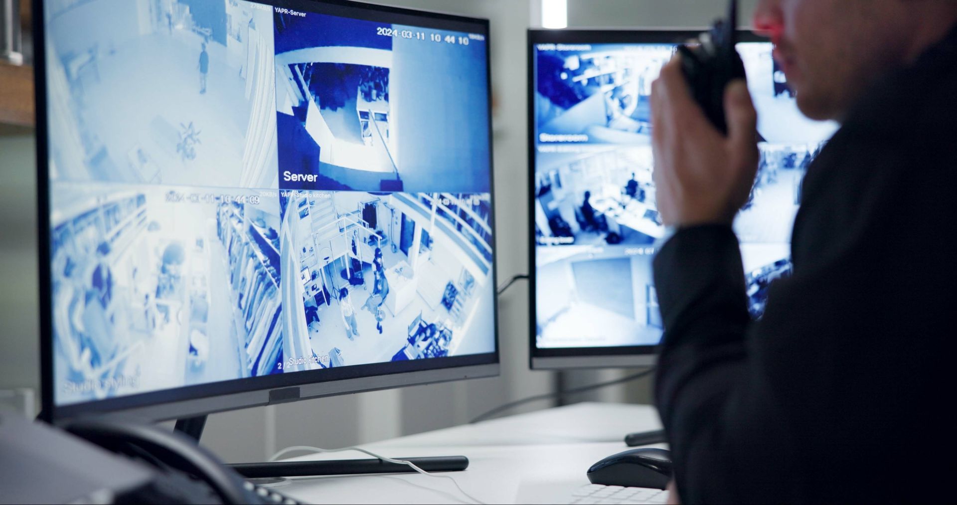 Security guard monitoring multiple camera feeds on two computer monitors while using a walkie-talkie.