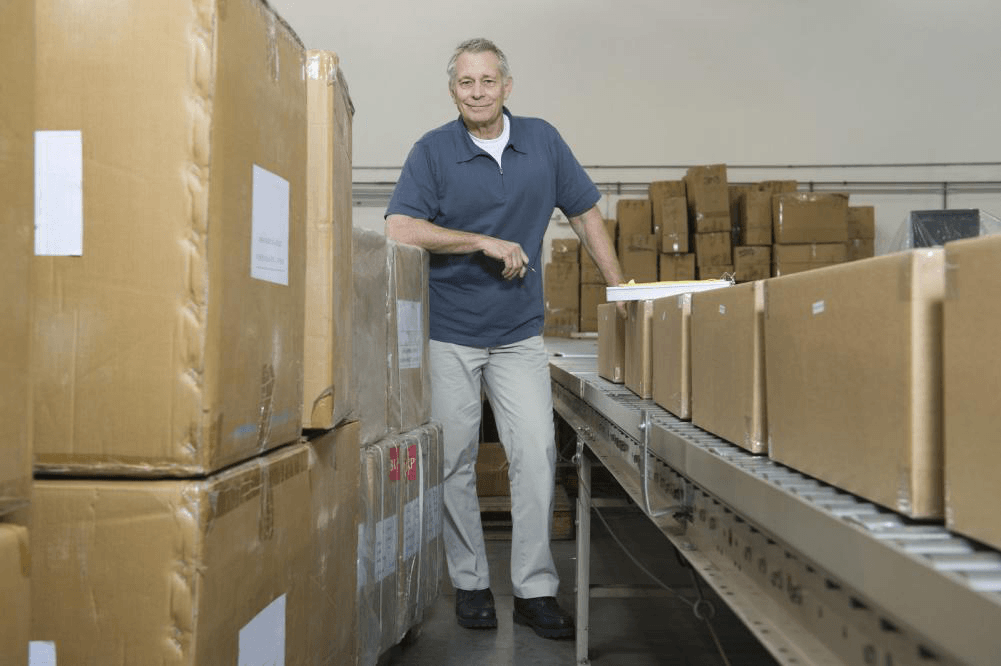 A man is standing next to a conveyor belt filled with boxes in a warehouse.