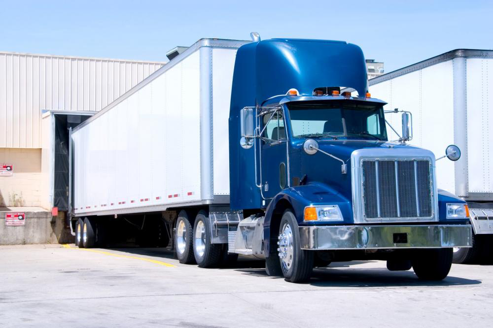 A blue semi truck with a white trailer is parked in front of a warehouse.