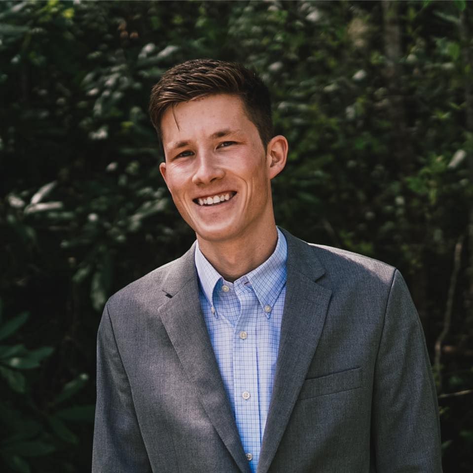 Smiling man in a gray blazer and blue shirt, posing outside in front of foliage.