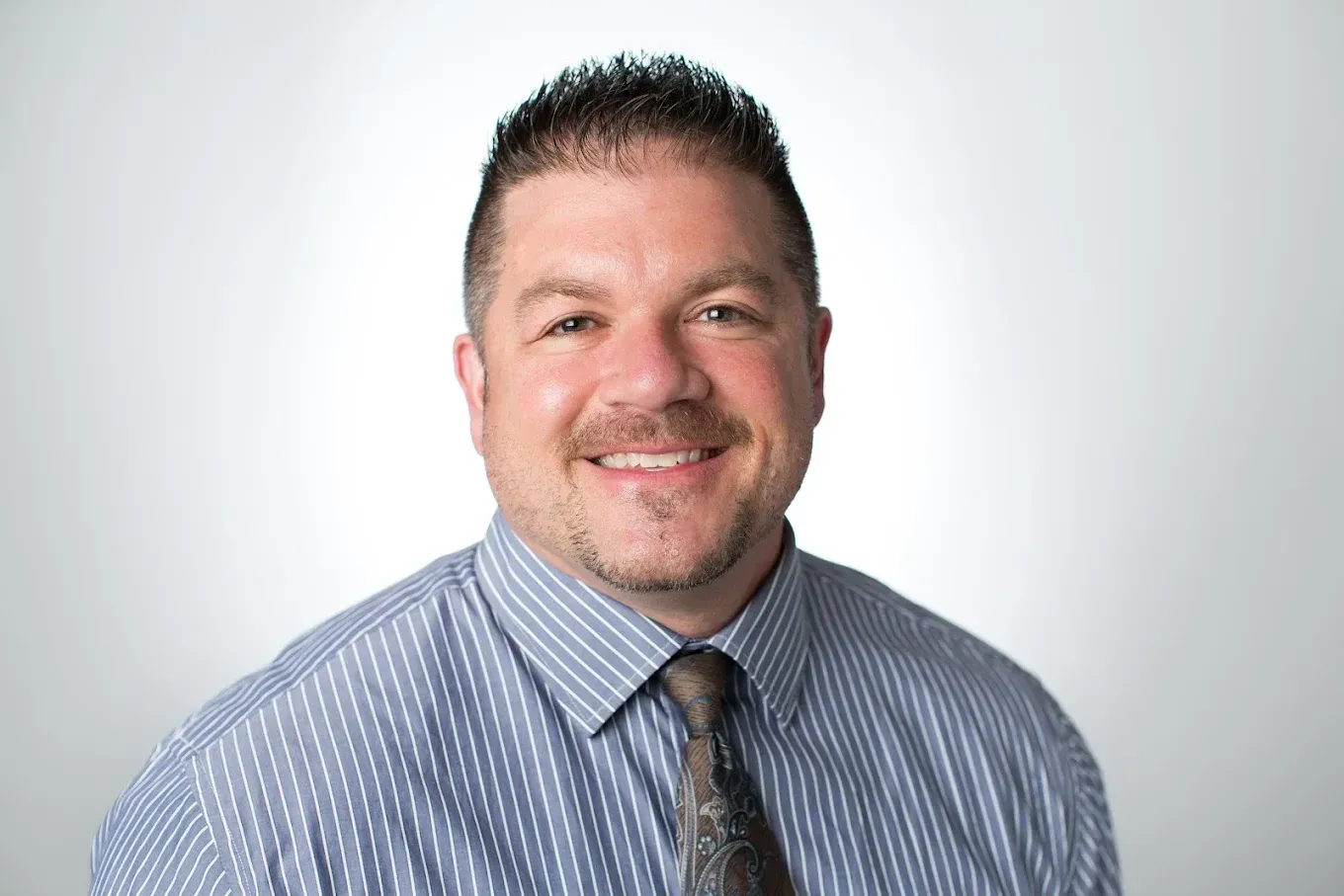 Man with dark hair, smiling in blue striped shirt and tie against a white background.