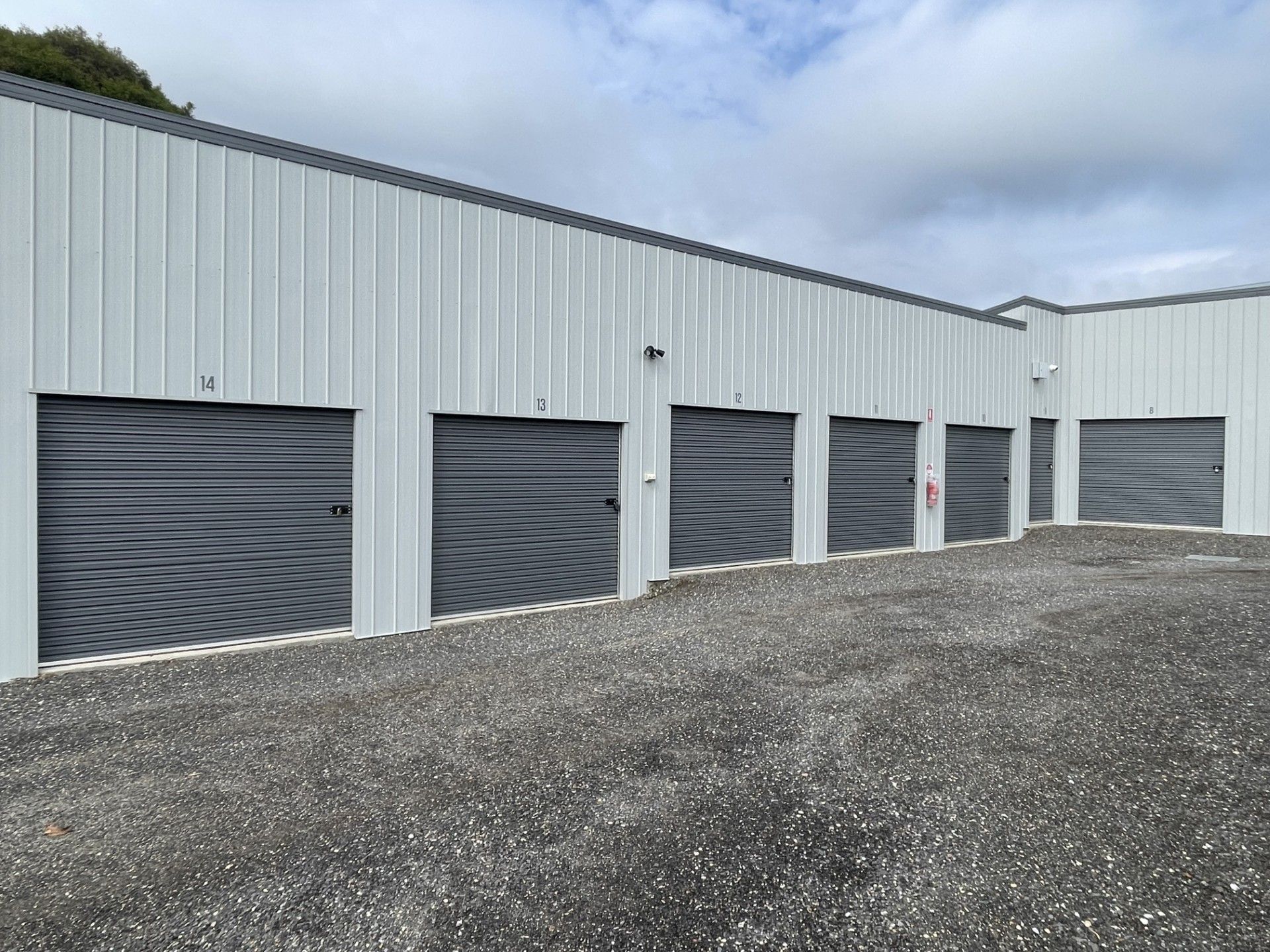 A row of grey metal self-storage units with roll-up doors, set against a cloudy sky over a gravel ground.