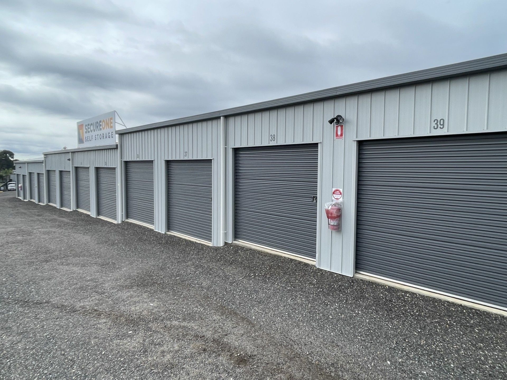 A row of gray storage units with closed roll-up doors, numbered on a gravel lot under an overcast sky.