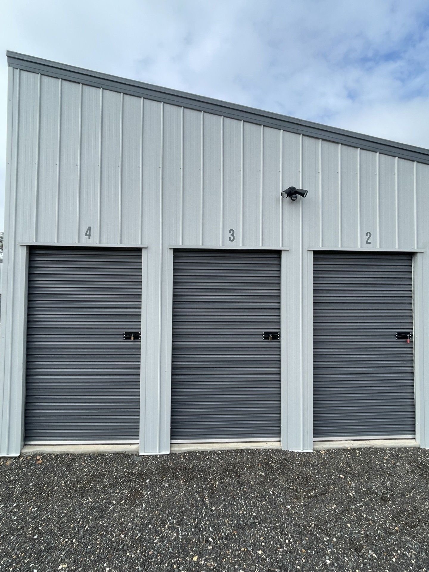 White storage building with three numbered grey roll-up doors, labeled 2, 3, and 4, situated on a gravel lot.