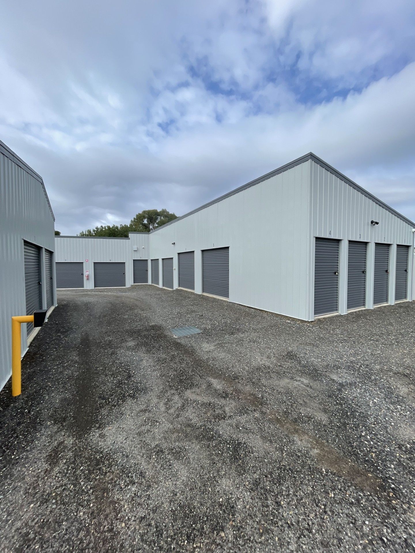 A gravel lot in front of white, metal-sided storage unit buildings under a cloudy, blue sky.
