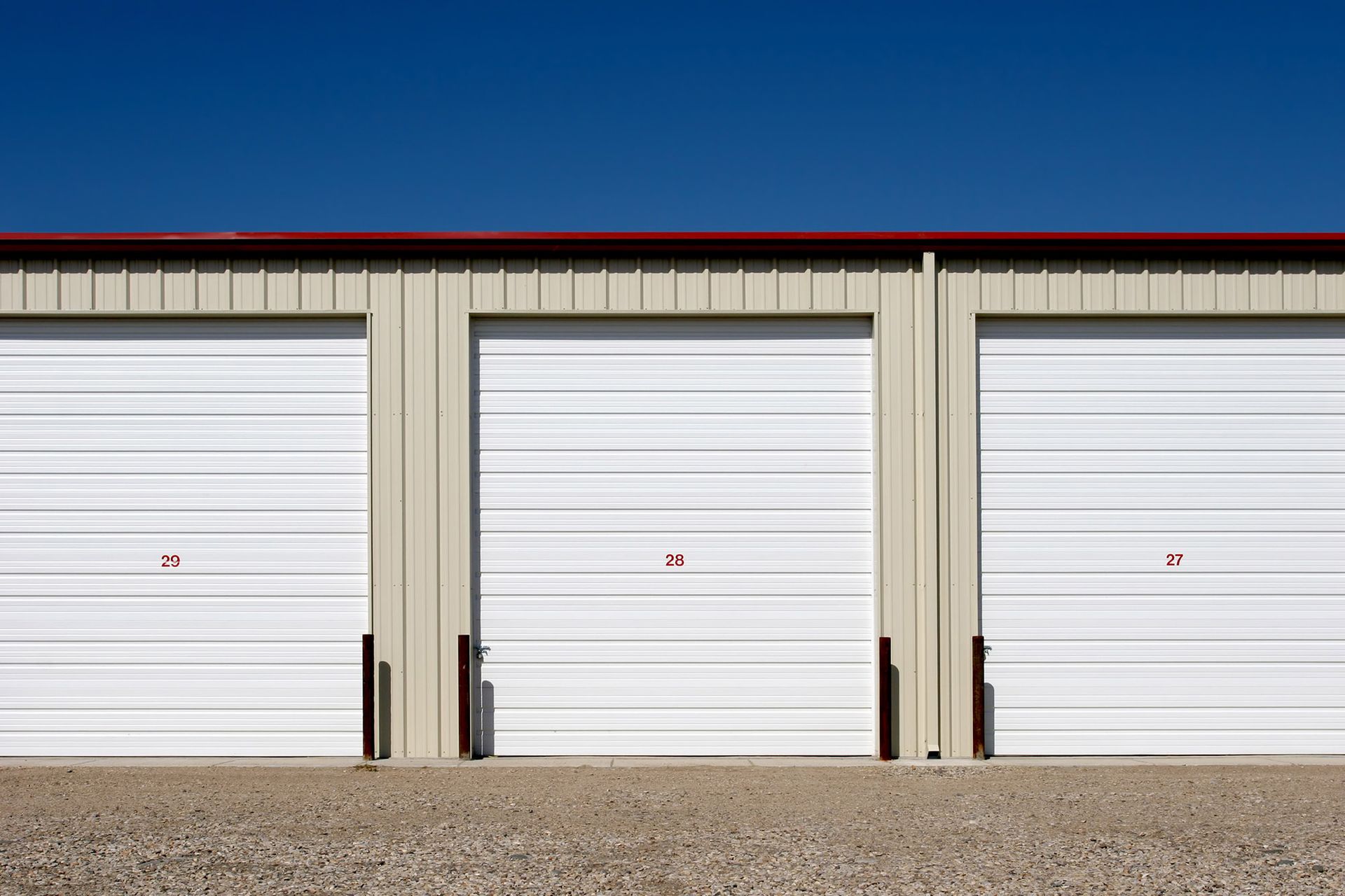 Three white storage unit doors with red numbering in a metal building against a blue sky.
