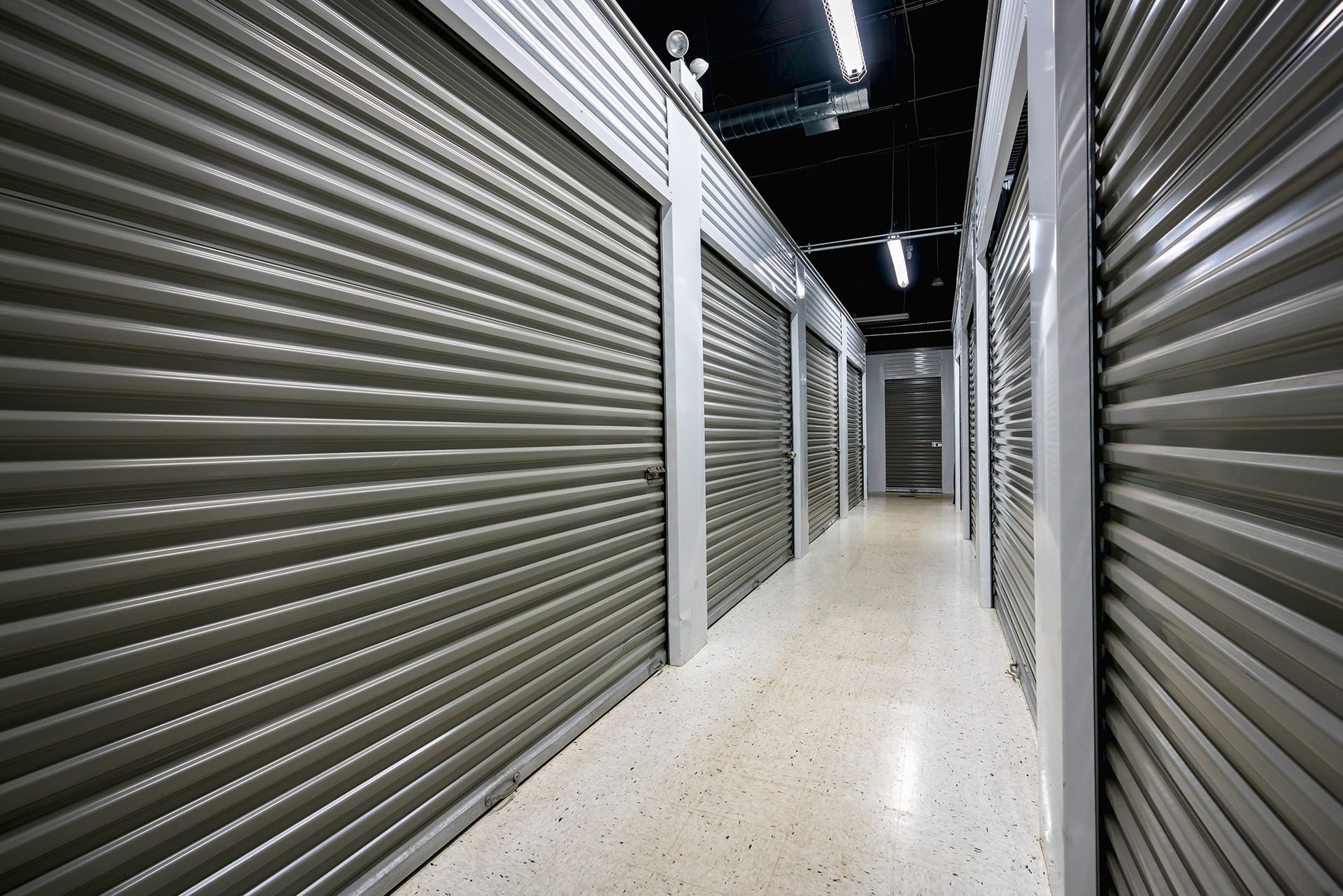 A narrow hallway inside a self-storage facility, lined with rows of closed gray metal roll-up doors on both sides.