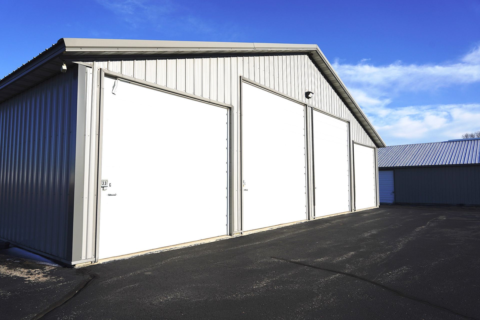 A light-gray metal storage building with three closed white garage doors sits on an asphalt lot under a blue sky.