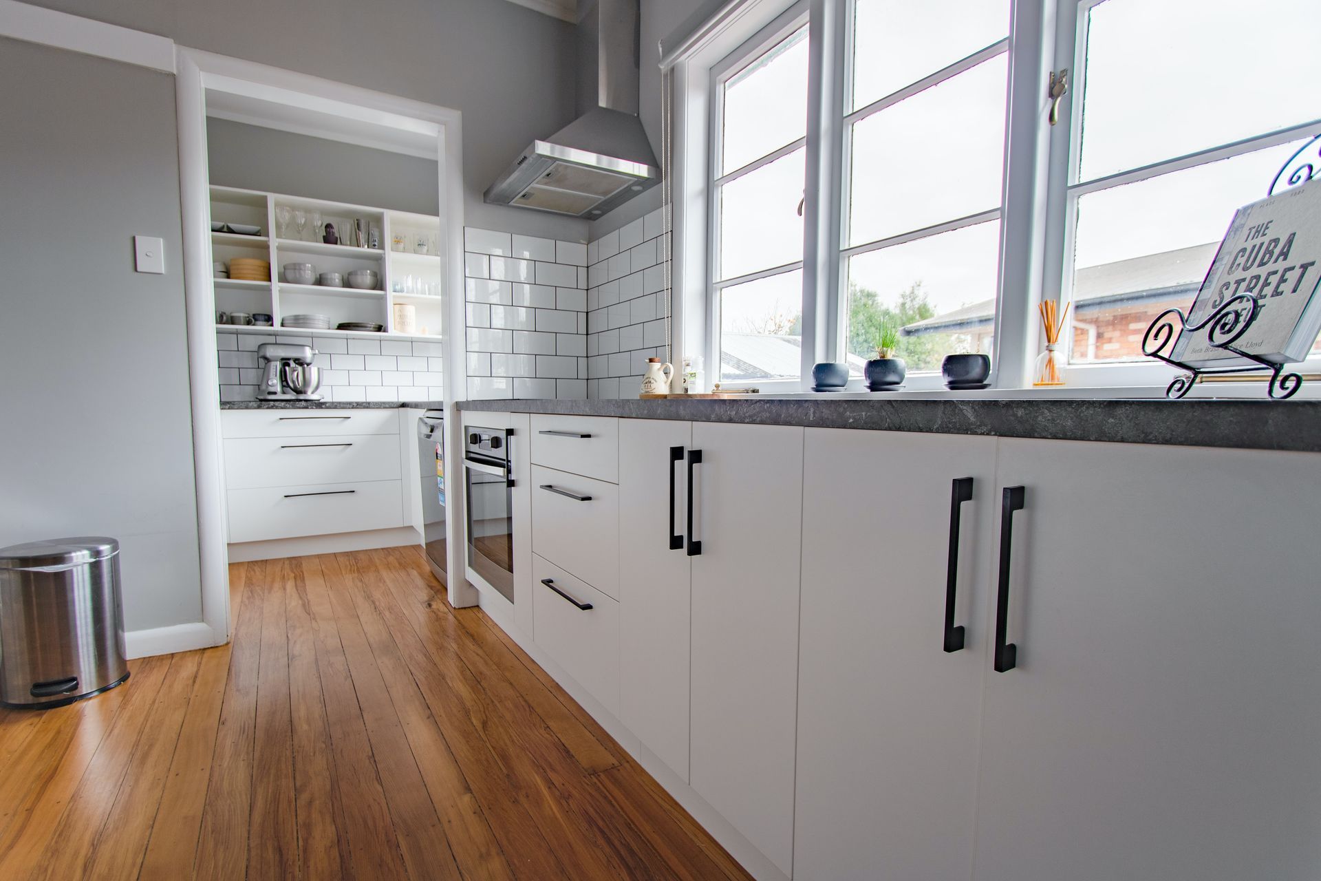 A kitchen with white cabinets and wooden floors and a large window.