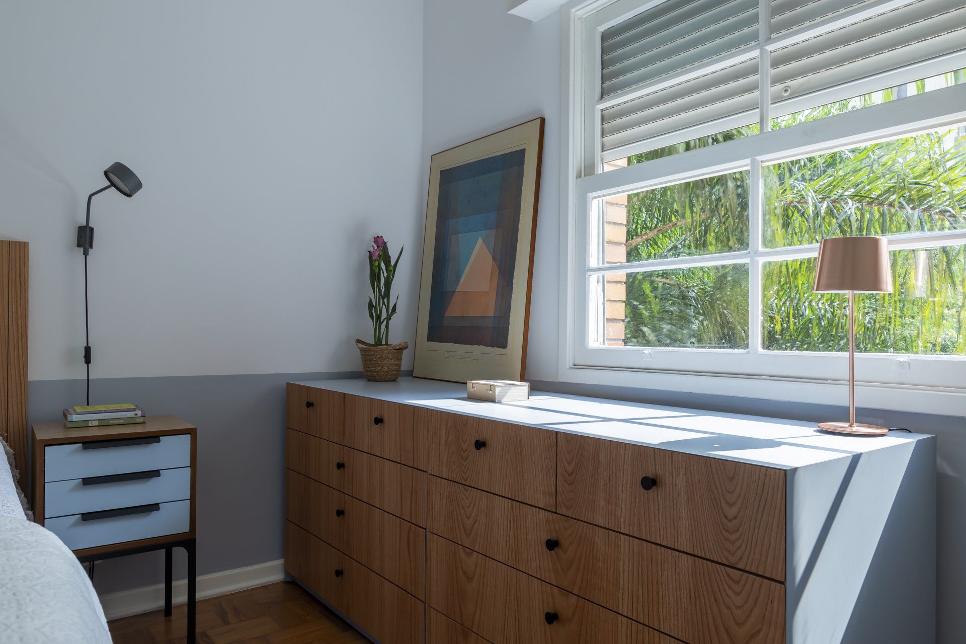 Um quarto com cómoda, mesa de cabeceira, candeeiro e janela.