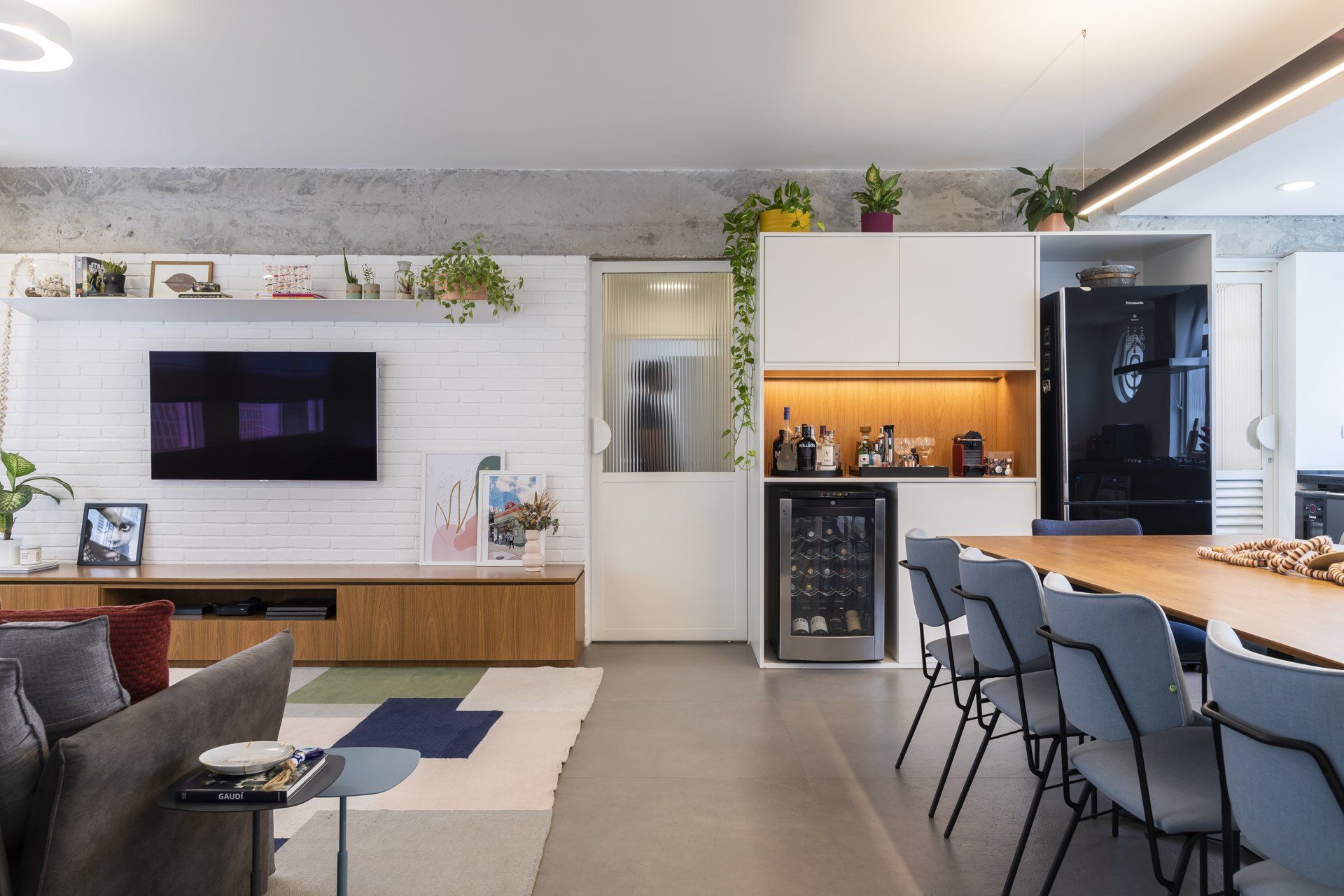Uma sala de estar com mesa de jantar, cadeiras, televisão e geladeira.